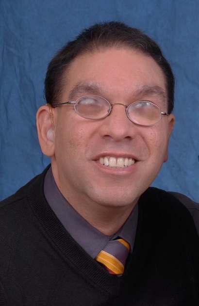 A Latino man with short brown hair in front of blue background